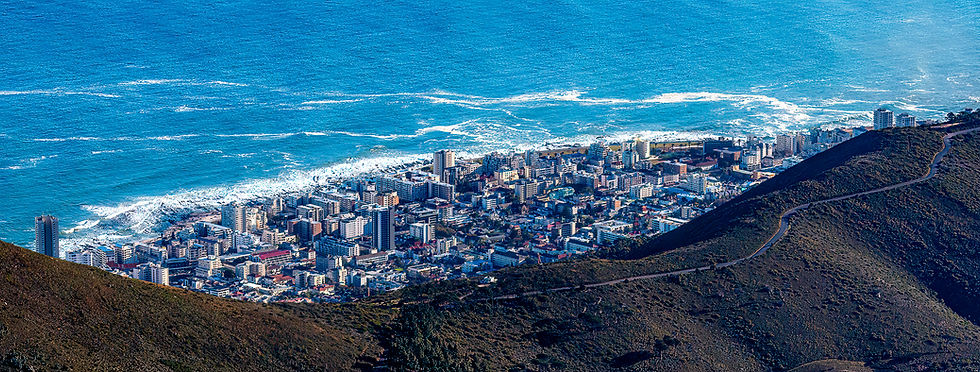 Aerial view of a city with skyscrapers by a blue ocean, surrounded by green hills. Waves crash on the shore under a clear blue sky.