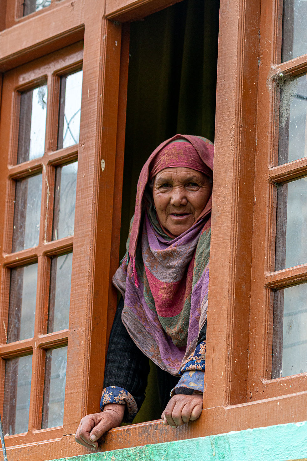 Elderly woman in colorful headscarf leans out a brown-framed window, smiling. Background shows green wall and wooden window details.