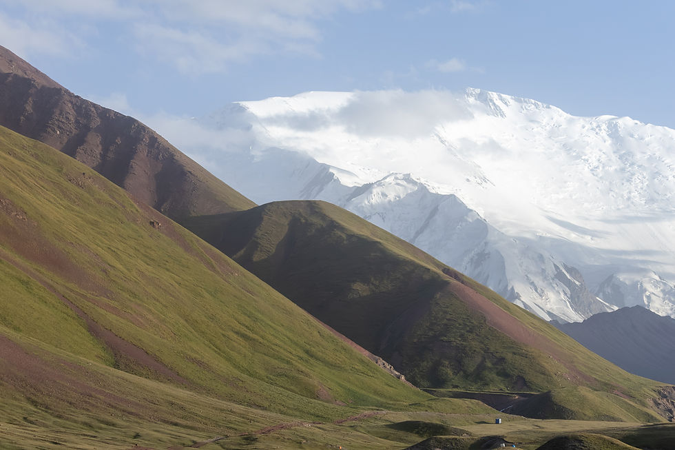 Green hills under a clear sky, with snow-capped mountains in the background. The scene is tranquil and expansive, with soft sunlight casting shadows.