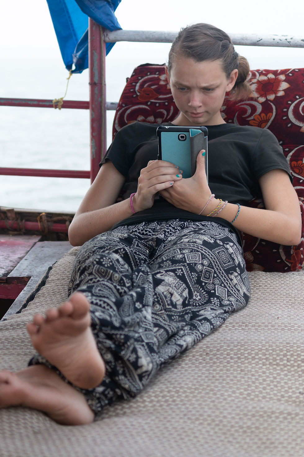 Teen girl lying on a patterned mat looks intently at a tablet. She's wearing patterned pants and a black shirt with the sea in the background.