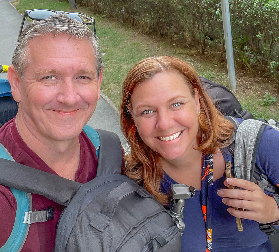Two smiling people with backpacks pose for a selfie outdoors. A pathway and green bushes are in the background. Sunny day.