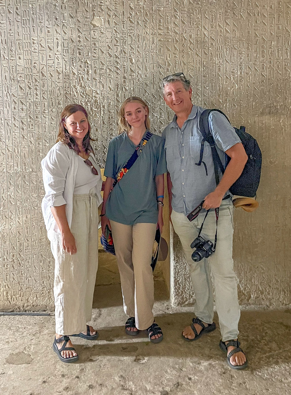Three people smiling in front of a wall with ancient hieroglyphs. They wear casual clothing and carry a camera. Stone floor visible.