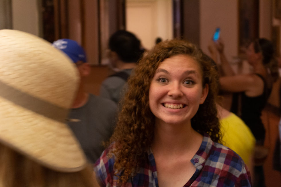 Young woman smiling in a plaid shirt in a crowded indoor space. Others are blurred in the background, one taking a photo with a phone.