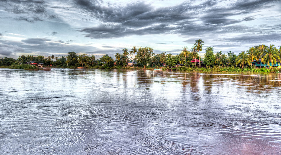 Calm river reflecting cloudy sky, with lush greenery and palm trees along the shoreline. Huts and red-roofed buildings are visible.