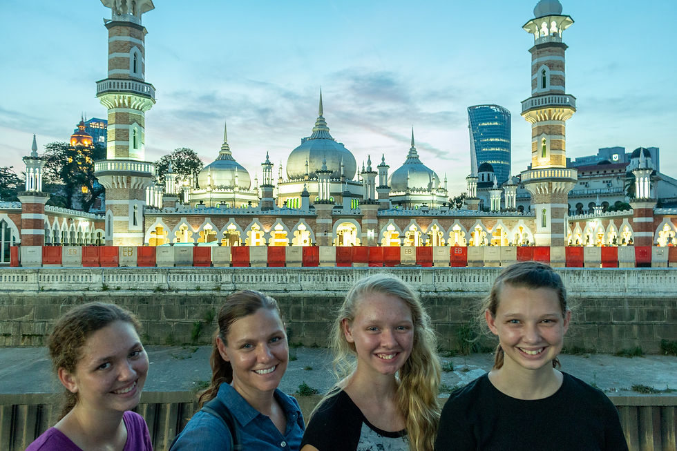 Four people smiling in front of an illuminated mosque with domes and minarets. The sky is a soft blue at dusk.