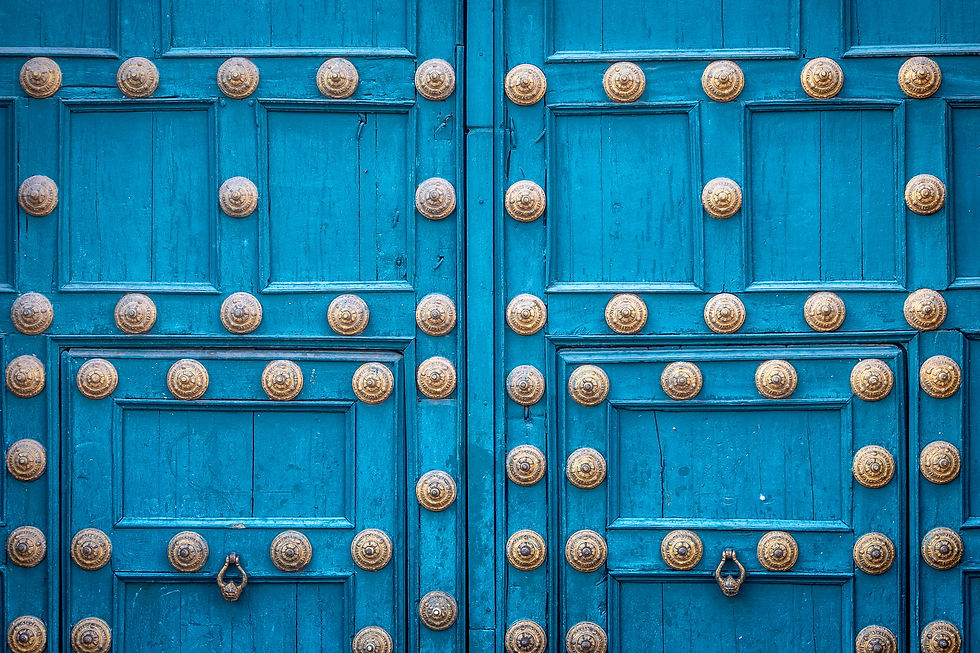 Blue wooden door with ornate gold studs and metal handles, featuring a symmetrical pattern. The texture is rustic, evoking a historic feel.