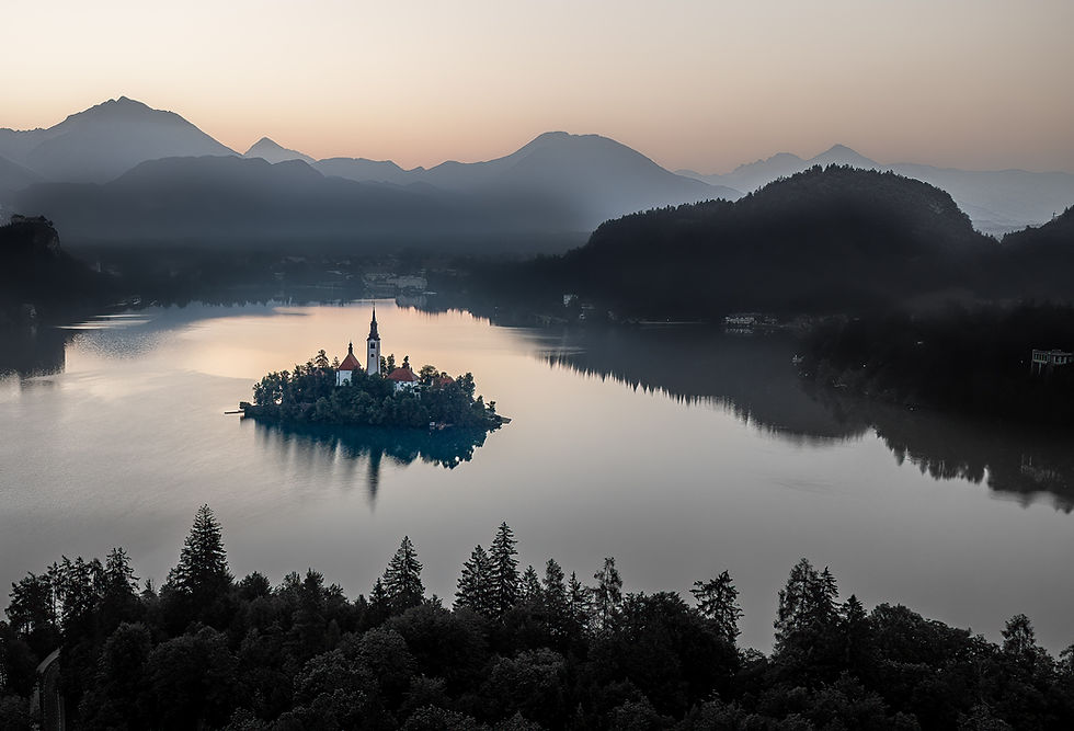 Island with a church in a serene lake at dawn, surrounded by misty mountains and silhouetted trees. Calm and peaceful atmosphere.