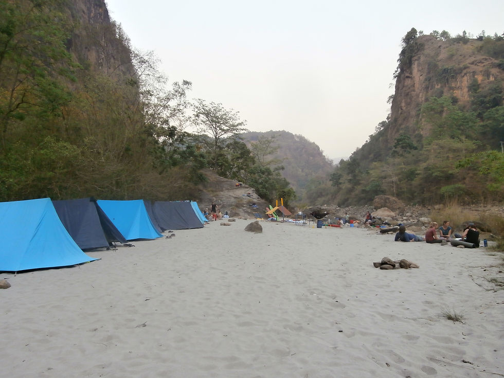 Blue tents on sandy ground in a canyon setting. People relax near campfires, surrounded by trees and cliffs under a calm sky.