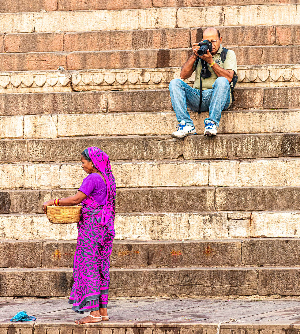 Man photographing a woman on stone steps. Woman in vibrant purple sari holds a basket, standing below. Rustic stone background.