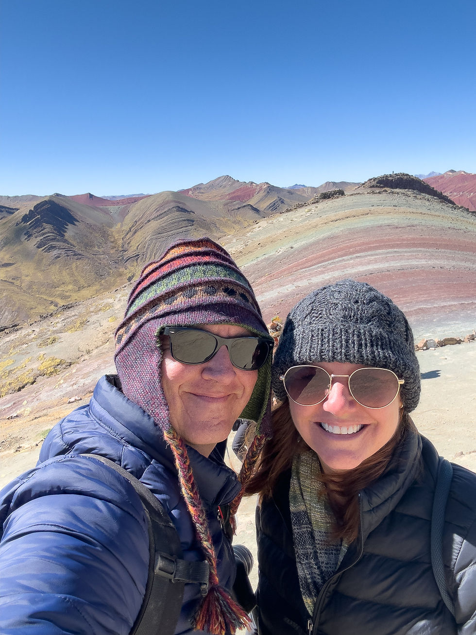 Two people in winter gear smile for a selfie at Rainbow Mountain, Peru. Colorful striped hat and sunglasses stand out against clear blue sky.