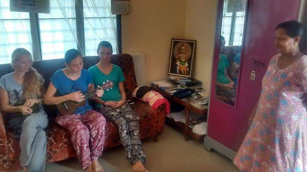 Three people sit on a sofa; one plays a ukulele. A woman stands nearby in a room with a purple wardrobe and a painting on the wall. Cozy setting.