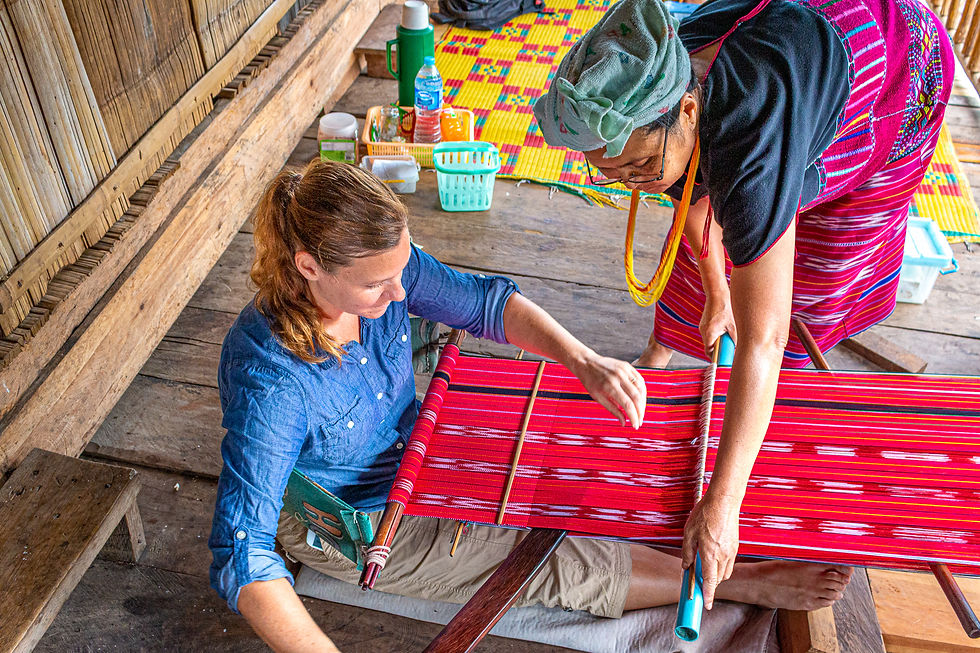 Two women weave on a red-patterned loom in a wooden setting. One guides the other, creating a collaborative and focused atmosphere.