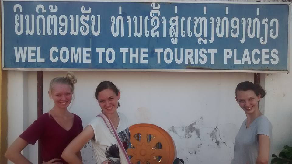Three people smile in front of a sign reading "WELCOME TO THE TOURIST PLACES" in blue and white. The setting is outdoors, sunny.