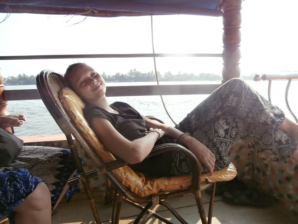 Person relaxing in a wicker chair on a boat, smiling with eyes closed. They wear patterned pants. River and trees visible in the background.