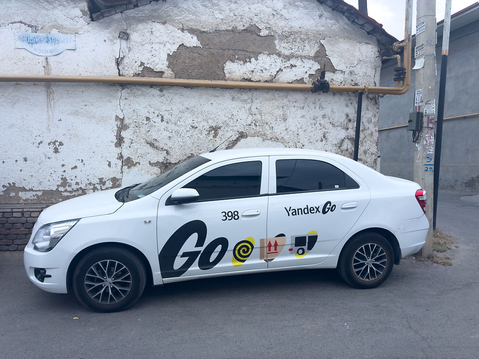White car with "Yandex Go" logo parked by a weathered wall. Number 398 and various icons visible. Urban setting, old building background.