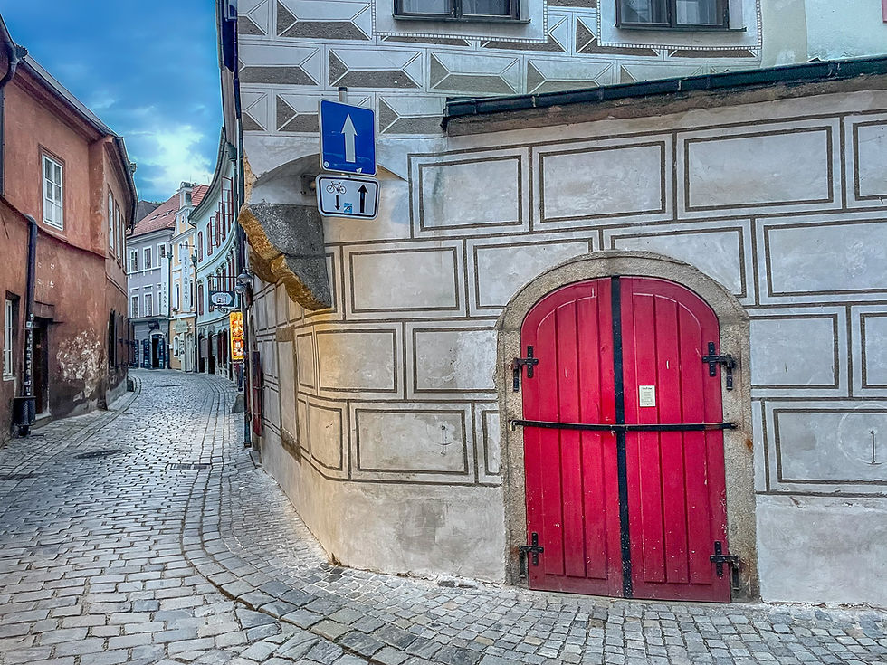 Narrow cobblestone street with colorful buildings, prominent red door with geometric patterns, and blue sky. Traffic signs visible. Calm mood.