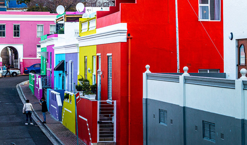 Colorful street with multicolored houses in pink, red, yellow, green, and blue. Two people walk on the sidewalk, creating a lively scene.