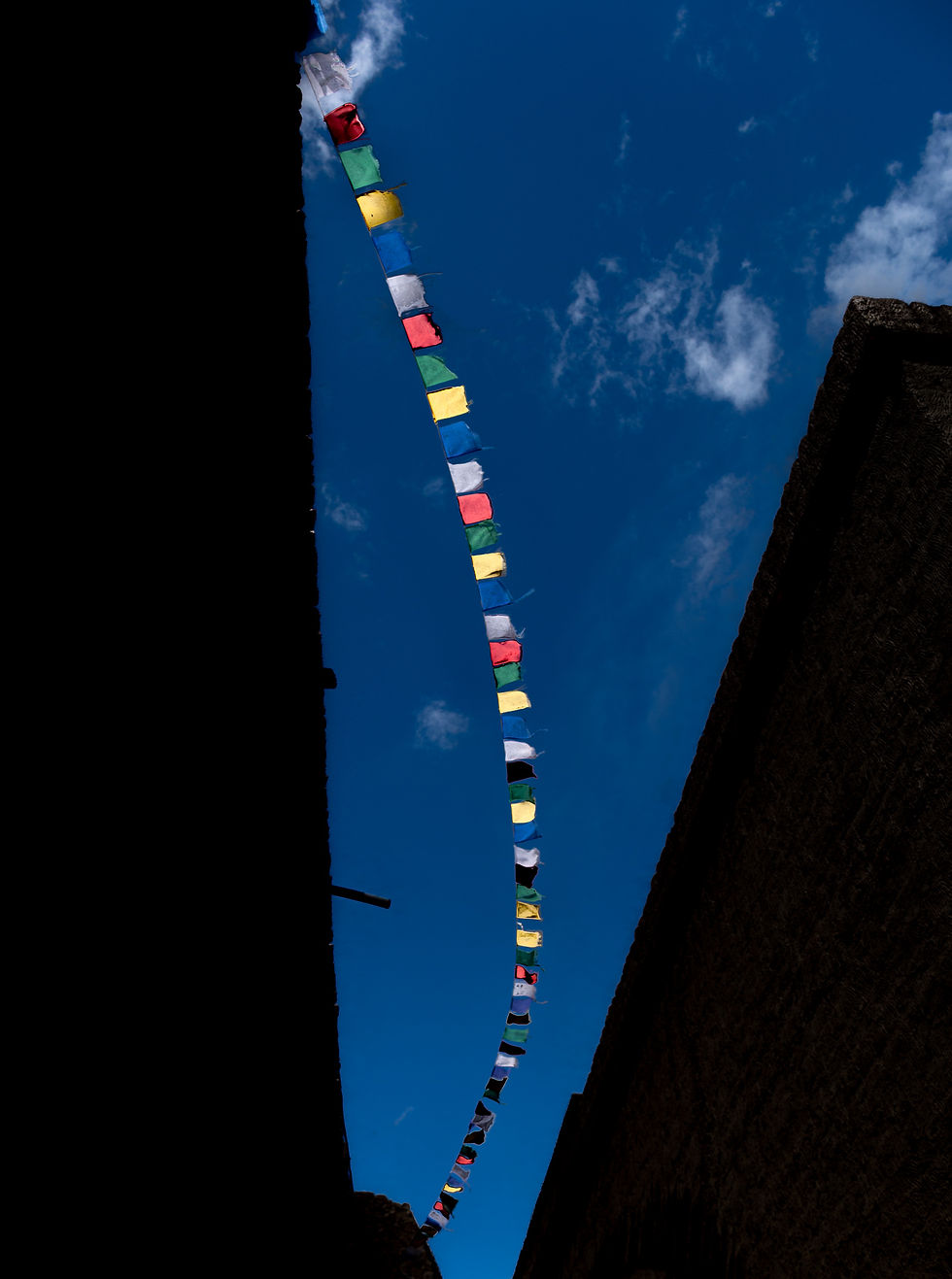 Colorful prayer flags strung between dark silhouettes of buildings, set against a bright blue sky with scattered clouds, creating a serene mood.