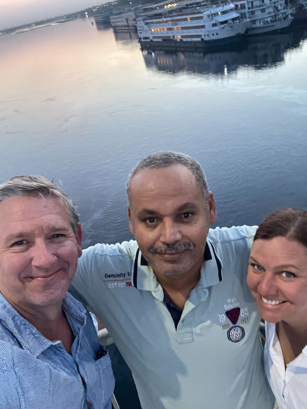 Three people smiling on a boat deck at sunset, with a calm river and bright cruise ship in the background. The mood is cheerful and relaxed.