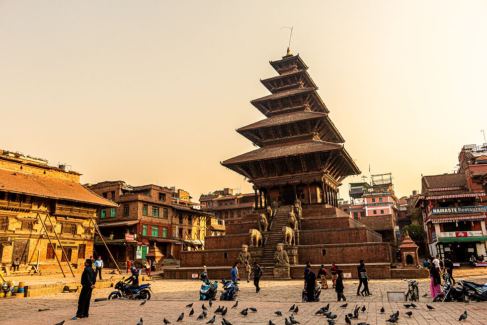 Ancient temple with tiered roof, surrounded by pigeons and people. Warm evening light, brick buildings, and a "Namaste Restaurant" sign.