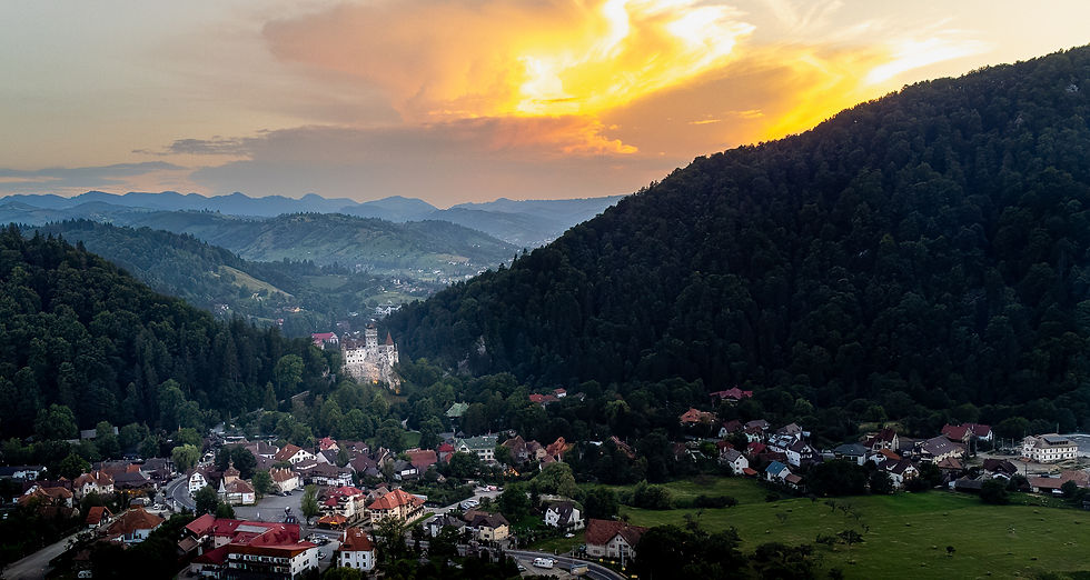 Aerial view of a quaint village surrounded by lush green forests and hills, with a dramatic orange sunset sky above a visible castle.