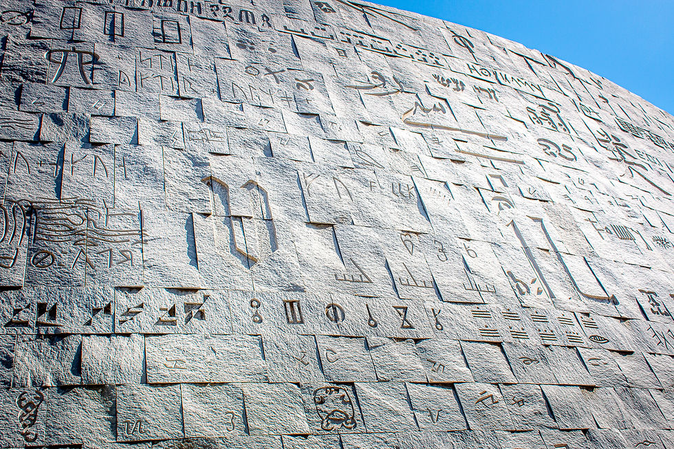 Wall with engraved symbols and scripts in a variety of languages under a clear blue sky. The stone surface creates a textured pattern.