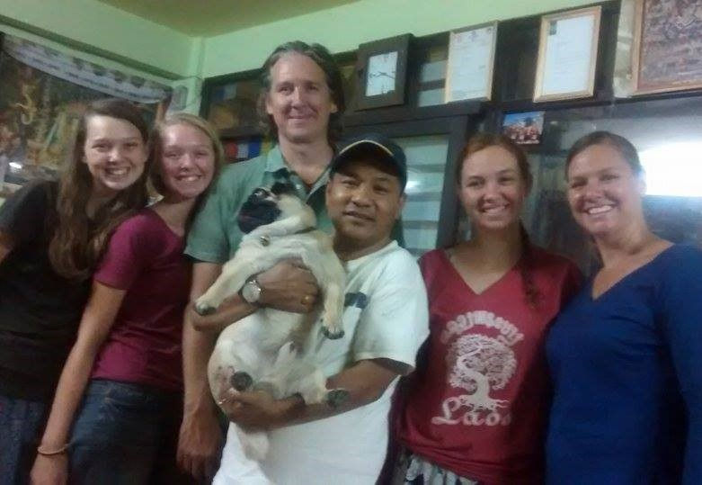 A group of six people smiles indoors. One person holds a pug. Green walls and framed photos are in the background. Casual, cheerful mood.