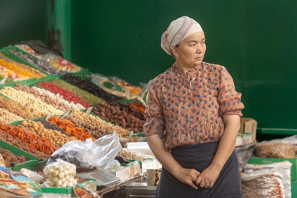 Woman at a market stall with a variety of dried fruits and nuts in colorful trays. She wears a patterned blouse and headscarf, looking pensive.