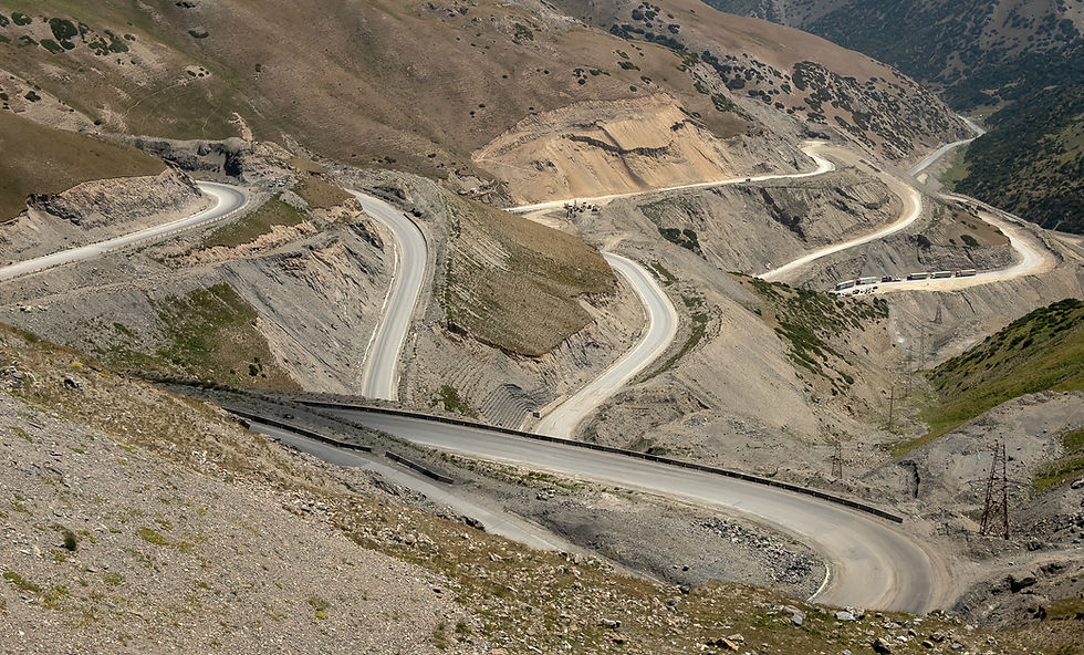 Winding mountain road with multiple hairpin turns, surrounded by arid, rocky hills. Sparse greenery and a clear sky in the background.