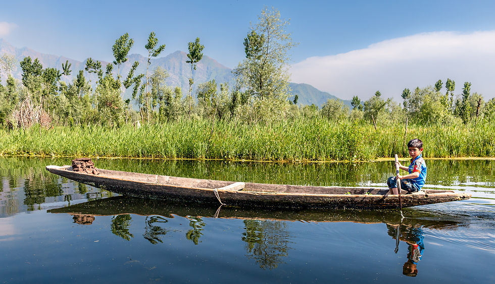 Child paddles a wooden boat on a tranquil river, surrounded by lush greenery and distant mountains under a clear blue sky. Reflective water.
