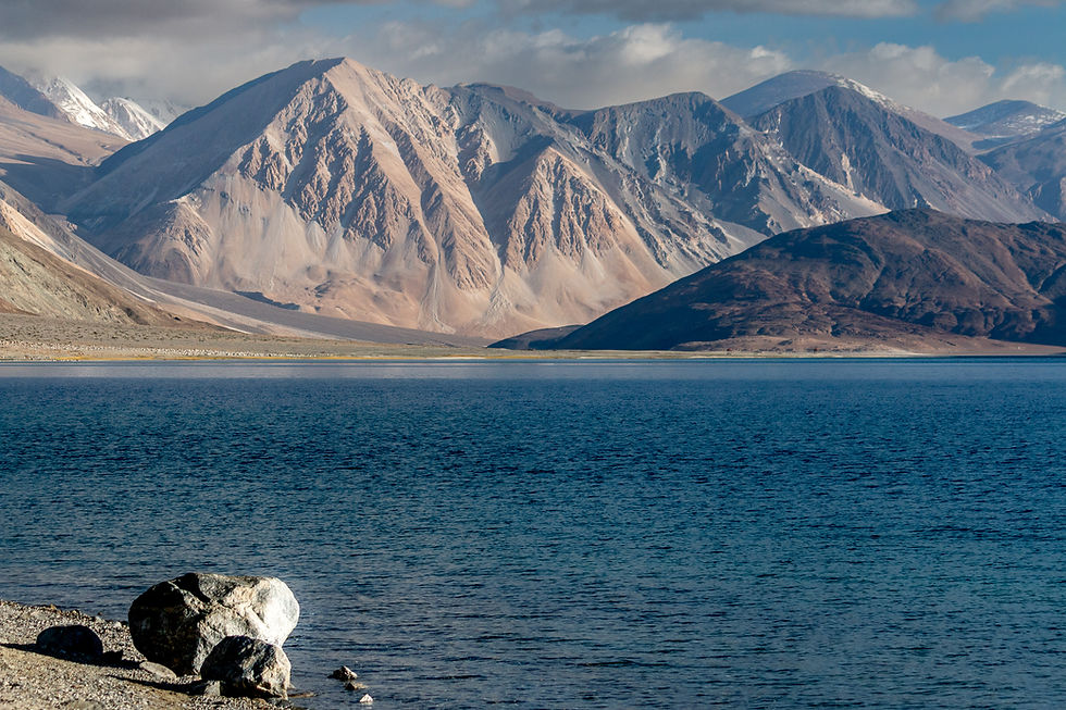 Rocky Pangong Lake lakeshore with a large boulder in the foreground, leading to a deep blue lake. Majestic, rugged mountains rise in the background.