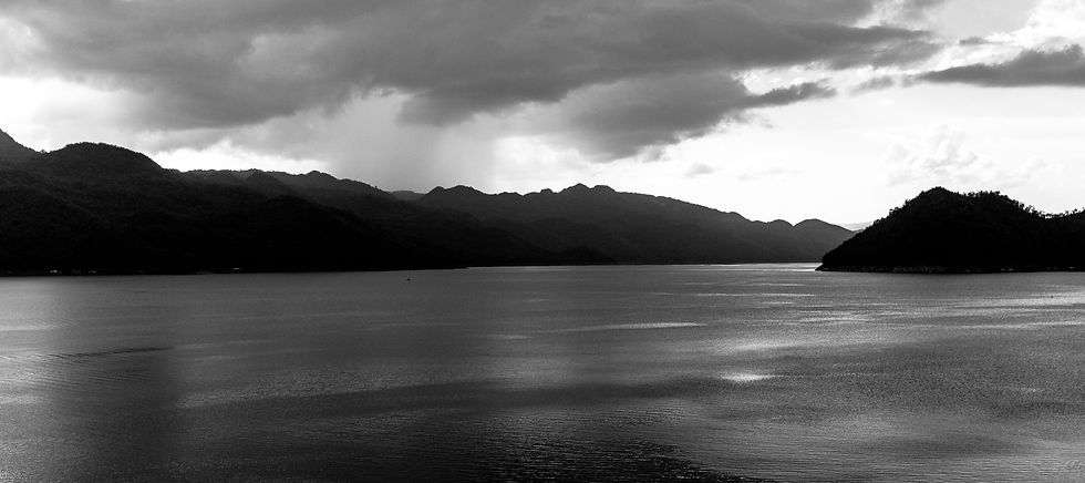 Calm waters stretch to distant hills under a moody, cloudy sky. The grayscale tones create a serene and contemplative atmosphere.