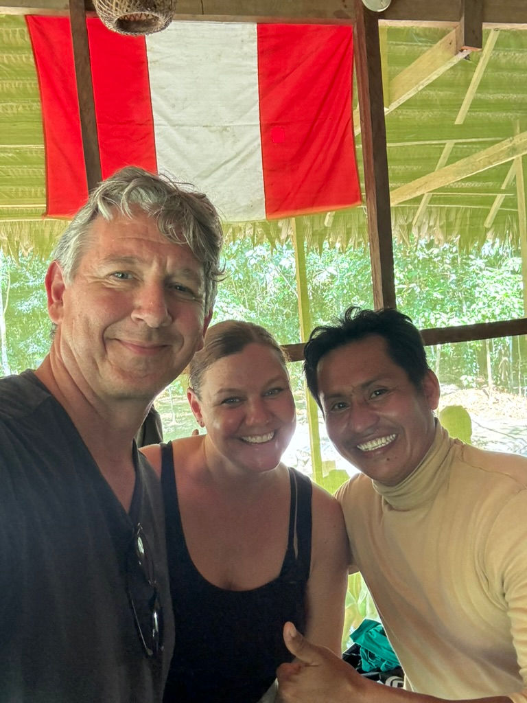 Three people smiling indoors with a red and white flag in the background. Natural light filters through the leafy surroundings.