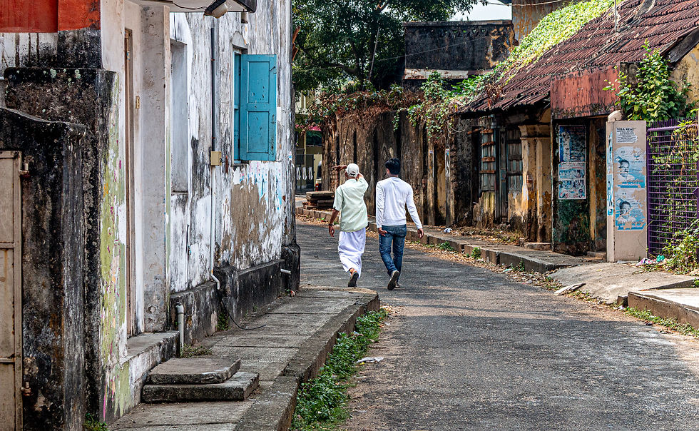 Two people walk down a narrow, weathered street with old buildings. One wears white, the other black and white. A blue window stands out.