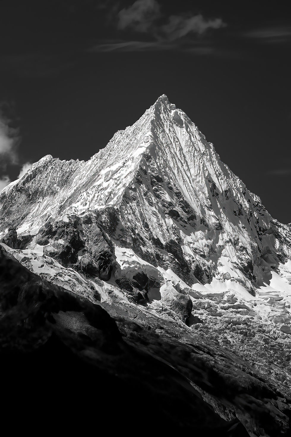 Snow-covered mountain peak under a dark sky, illuminated in light, with patches of shadow accentuating its rugged texture.