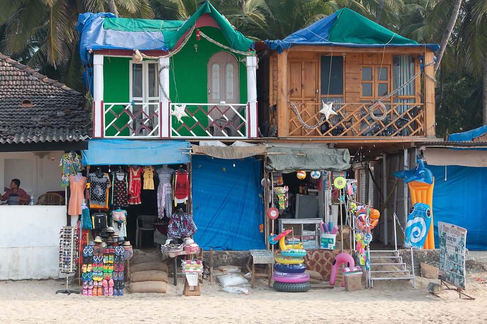 Beachside shops with colorful clothes, inflatables, and souvenirs under palm trees. Vibrant green and wooden huts above, relaxed atmosphere.