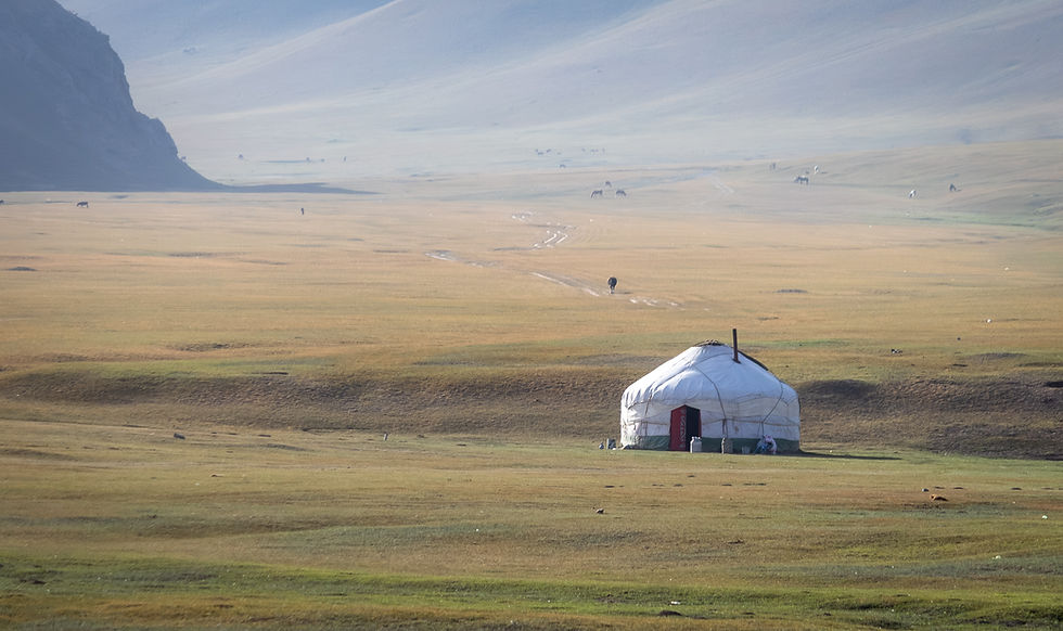 A yurt stands alone in a vast, open grassland with distant mountains. Grazing animals dot the landscape under a clear, blue sky.
