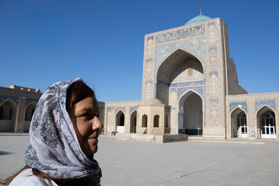 Woman in a patterned headscarf looks away, standing in front of a blue-tiled historical building under a clear blue sky.