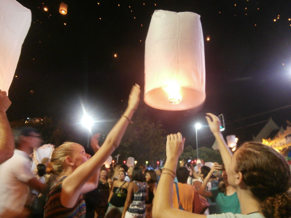 Crowd releases glowing paper lanterns into night sky. People reach upwards, illuminated by streetlights. Festive, joyful atmosphere.
