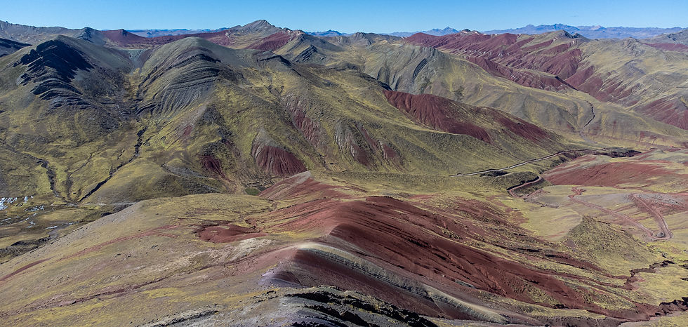 Aerial view of rolling mountains with red and green hues under a clear blue sky, creating a vibrant, serene landscape with winding paths.