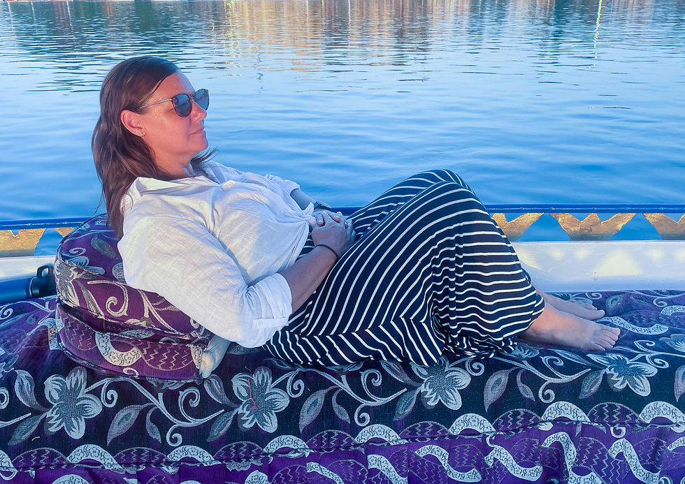 Woman relaxing on a patterned cushion on a boat, wearing a white shirt and striped skirt. Calm water in the background, serene mood.