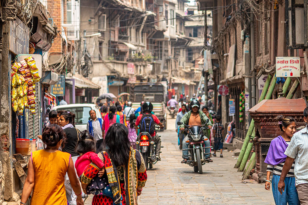 Busy street scene with people walking and motorbikes passing by. Shops line the road, bags of snacks hang outside. A lively, colorful urban atmosphere.