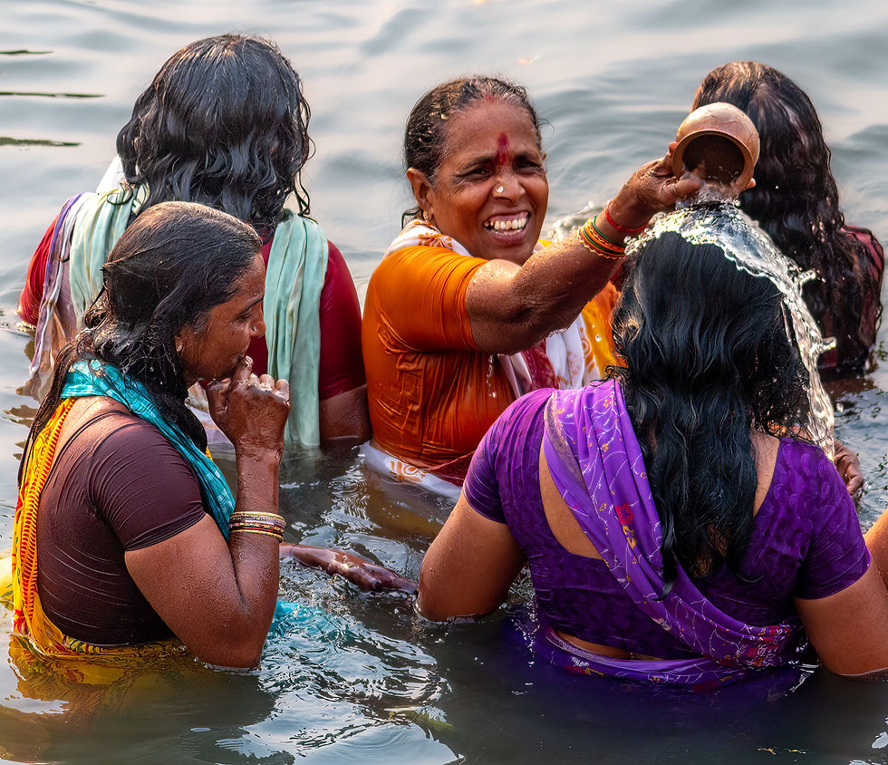 Women in colorful saris perform a joyful ritual in the water, smiling and pouring water from a pot. Sunlight reflects on the rippling surface.