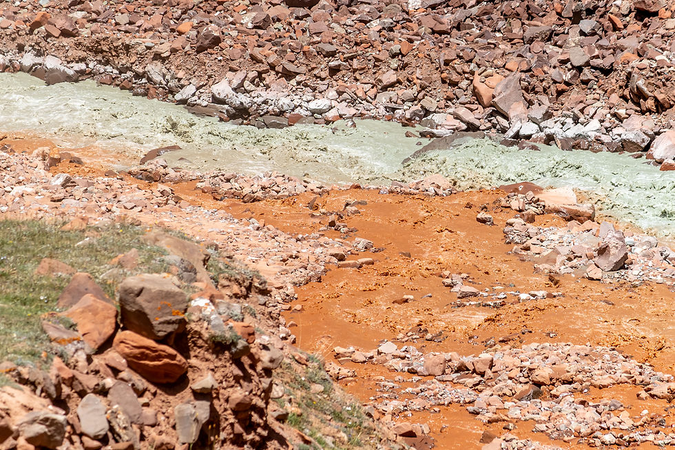 Rocky landscape with red and greenish streams flowing among scattered rocks. Earthy tones dominate the arid terrain.