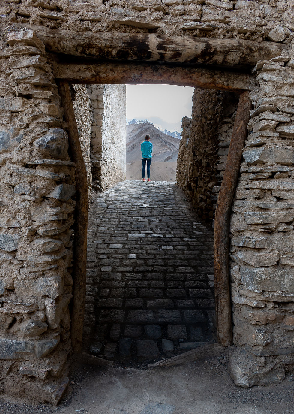 Person in blue jacket and red shoes stands in a stone archway, gazing at distant mountains. Rugged stone walls form a narrow passage.