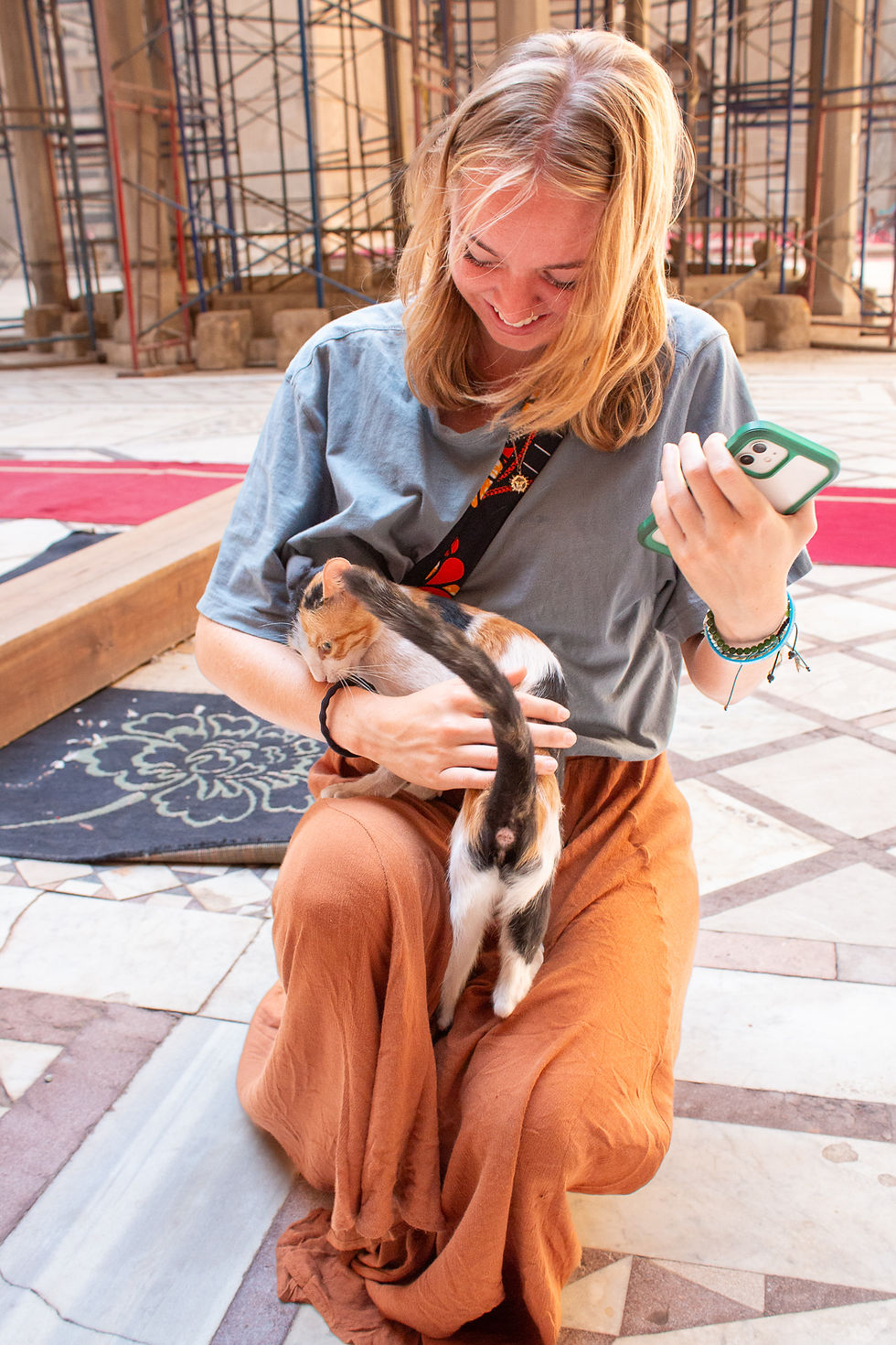 Woman kneels, smiling at a calico kitten in her lap inside a building with scaffolding. She holds a phone, wearing a blue shirt and orange pants.