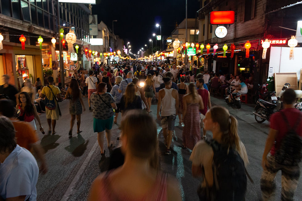Crowded night market street with people walking under colorful lanterns. Bright shop signs and lively atmosphere fill the scene.