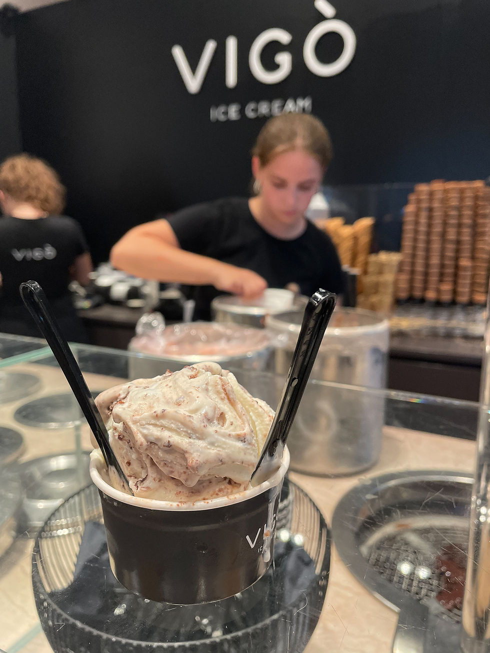 Ice cream in a black cup with two spoons on a counter. Two workers in black shirts scoop behind. "Vigò Ice Cream" text on wall.