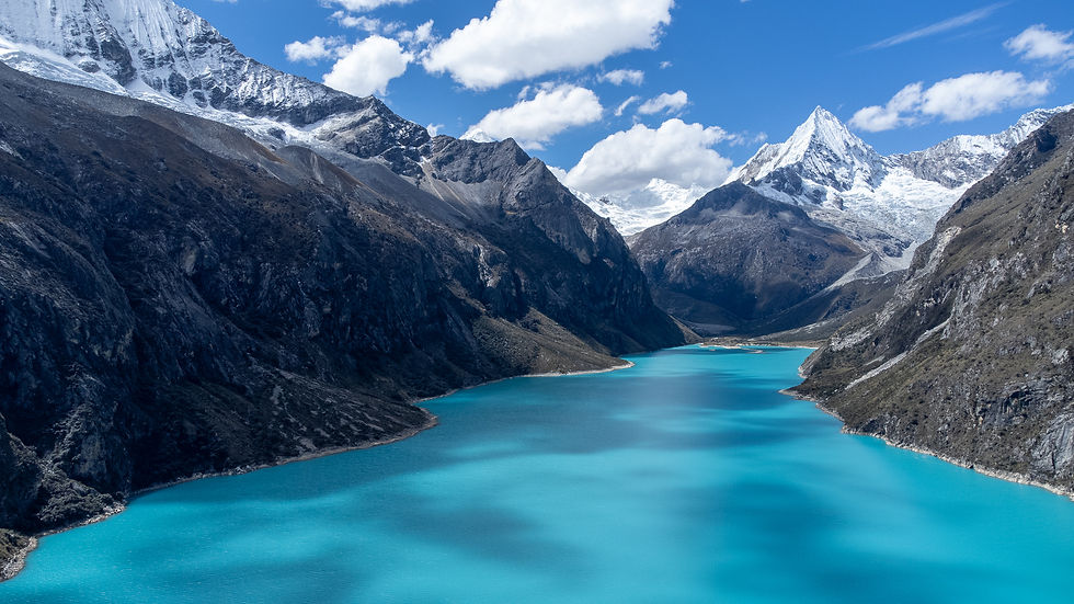 Turquoise lake surrounded by jagged mountains with snowy peaks under a bright blue sky and fluffy clouds. Serene and majestic landscape.