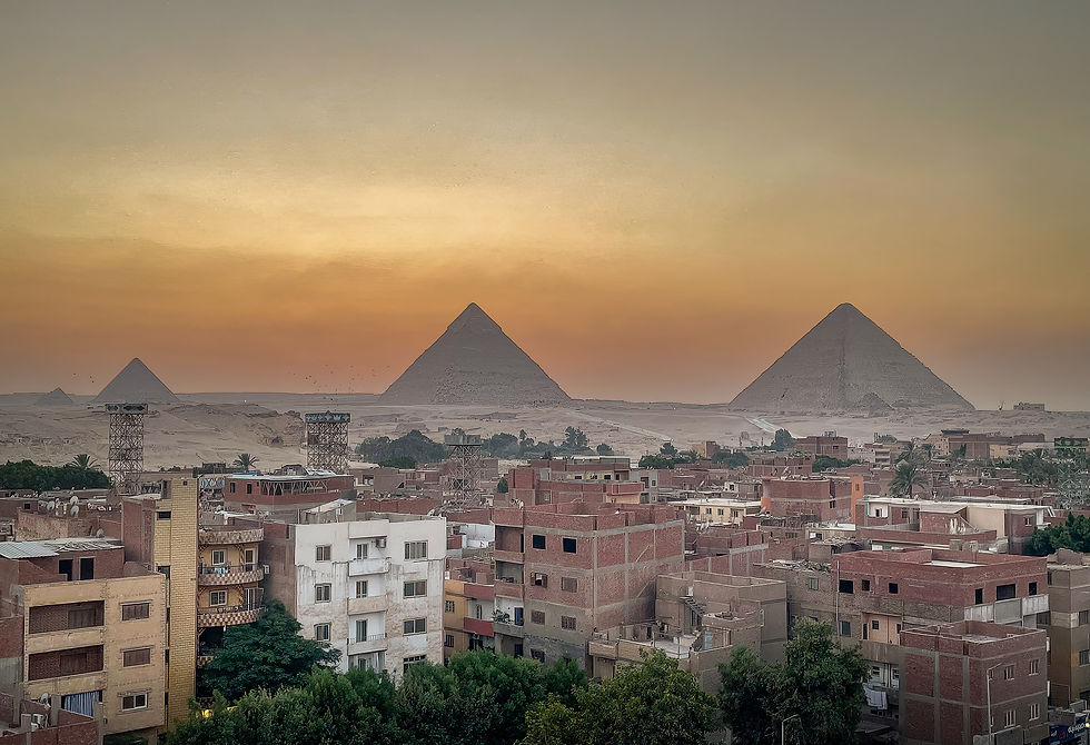 Sunset view of the pyramids near Giza, with rustic buildings in the foreground. Orange sky highlights the iconic structures. Calm mood.